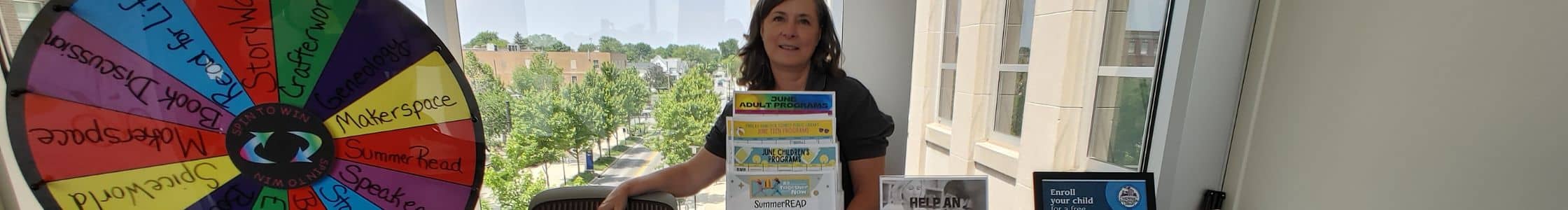 Outreach coordinator at an event with table of handouts of library information and colorful wheel for game